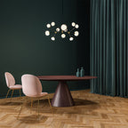 Glowing Clérachic chandelier in black and brass over a round dark wood table with pink chairs, against a deep green wall and curtain.