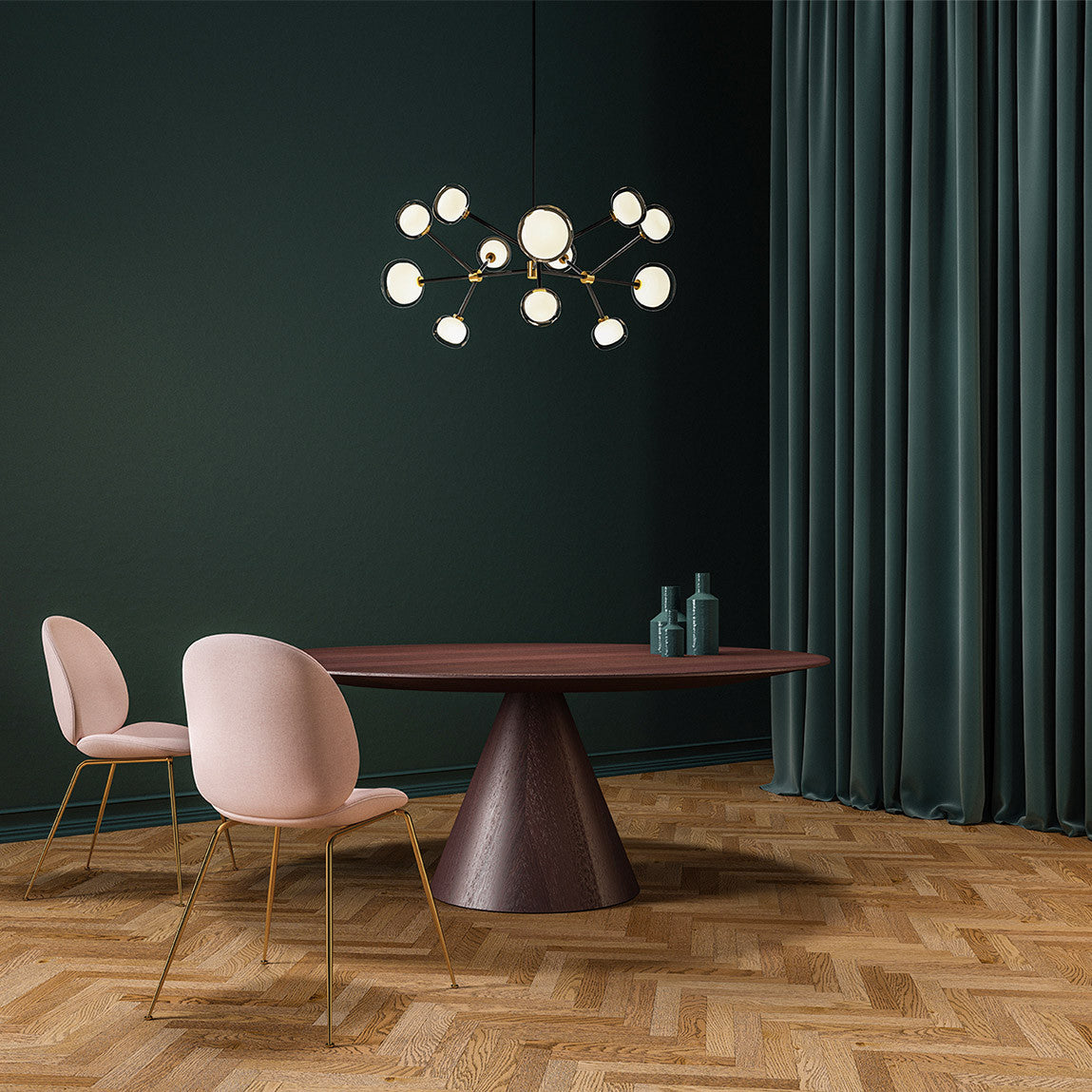 Glowing Clérachic chandelier in black and brass over a round dark wood table with pink chairs, against a deep green wall and curtain.
