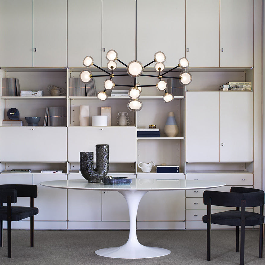 Lit Clérachic chandelier with black arms and round glass globes in front of cream storage cabinets, above a white oval table and black chairs.