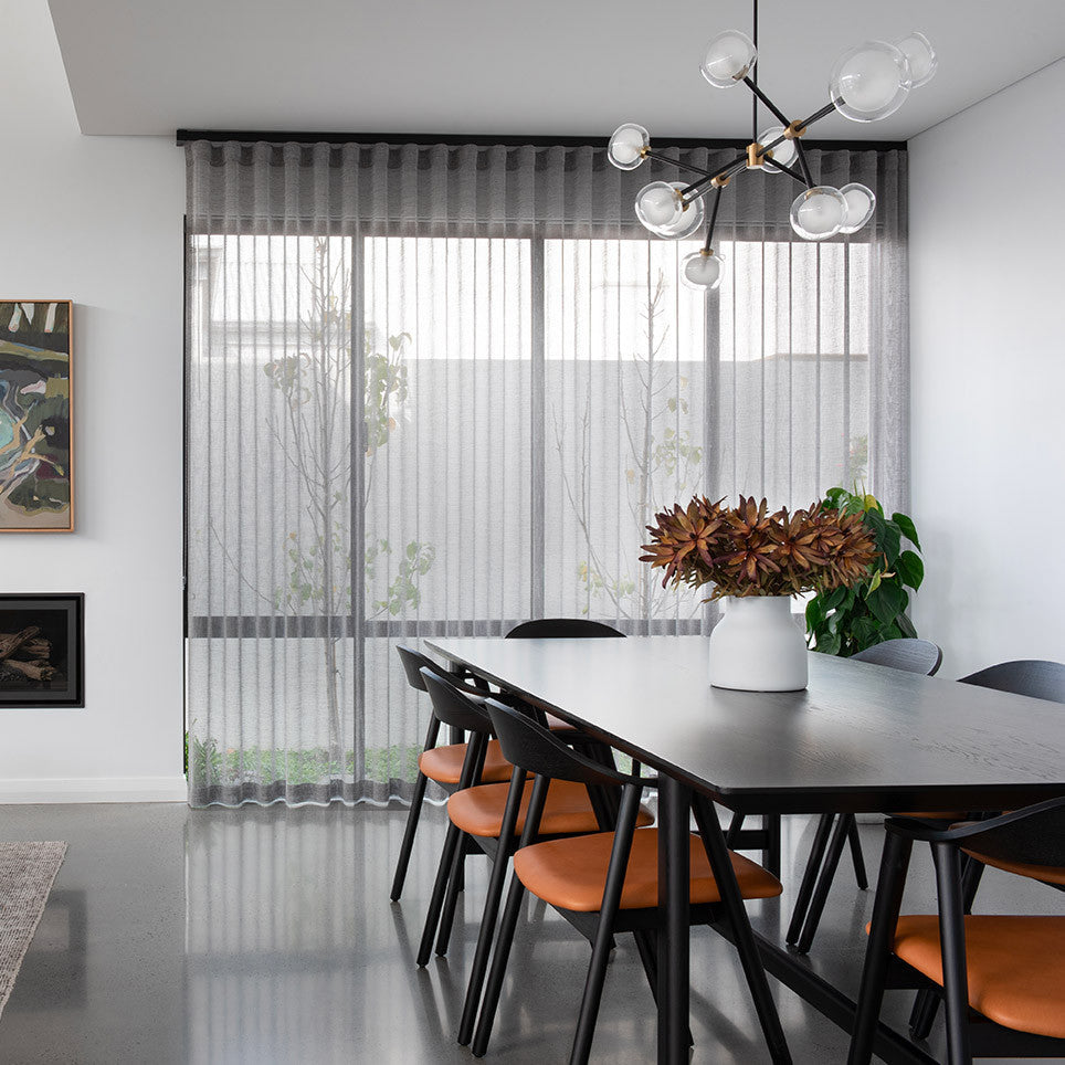 Clérachic chandelier with clear glass globes on black arms, above a dark dining table with orange-seated chairs and a grey sheer curtain.