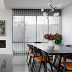 Clérachic chandelier with clear glass globes on black arms, above a dark dining table with orange-seated chairs and a grey sheer curtain.