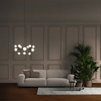 A lit Clérachic chandelier hangs in a panelled room above a grey sofa, with a tall green plant and a small side table.