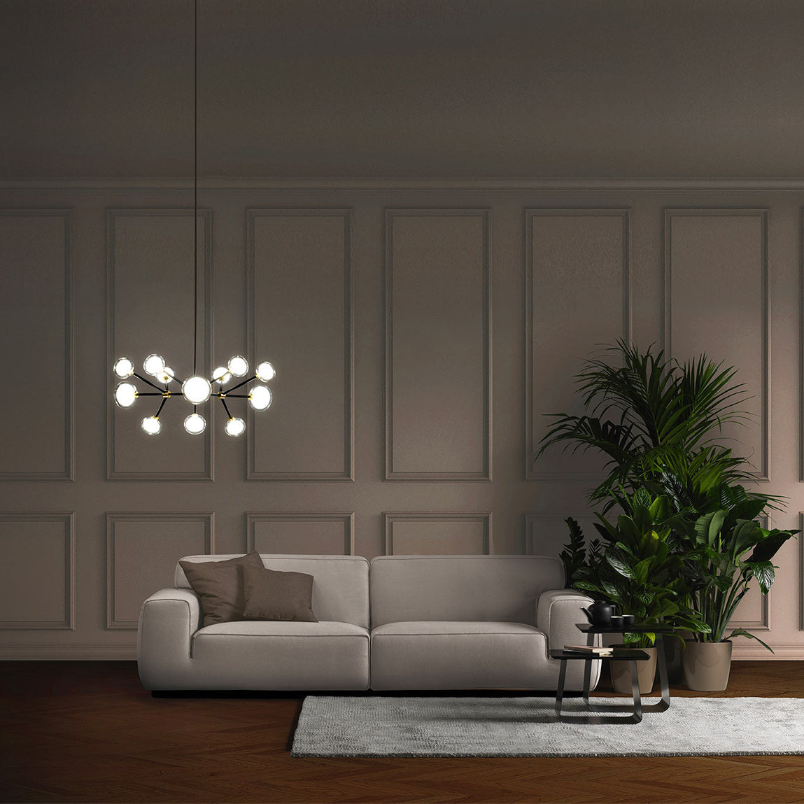 A lit Clérachic chandelier hangs in a panelled room above a grey sofa, with a tall green plant and a small side table.