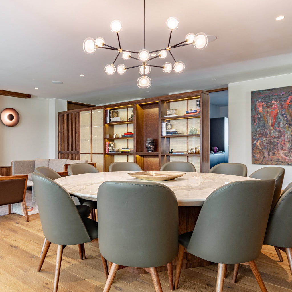 Lit Clérachic chandelier above a round marble dining table with grey-green leather chairs, in front of a walnut shelving wall.