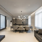 A Clérachic chandelier lit above a long marble dining table with grey chairs in a large, open room with a track light along the ceiling.