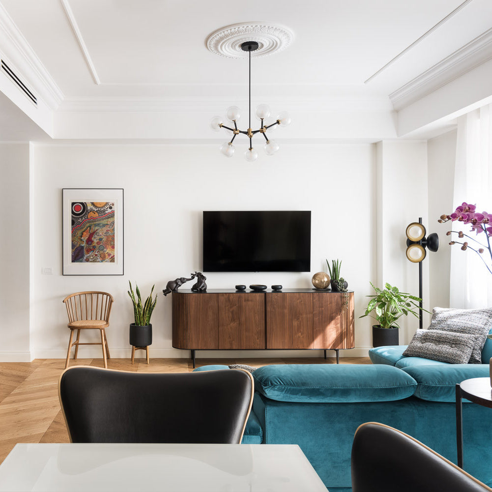Clérachic chandelier with clear globes beneath a ceiling rose, in a white living room with a walnut sideboard and a teal velvet sofa.