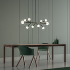 Lit Clérachic chandelier with a long black bar and glowing globes, above a walnut dining table with green chairs on a sage-green wall.