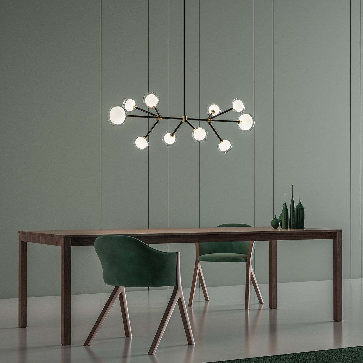 Lit Clérachic chandelier with a long black bar and glowing globes, above a walnut dining table with green chairs on a sage-green wall.