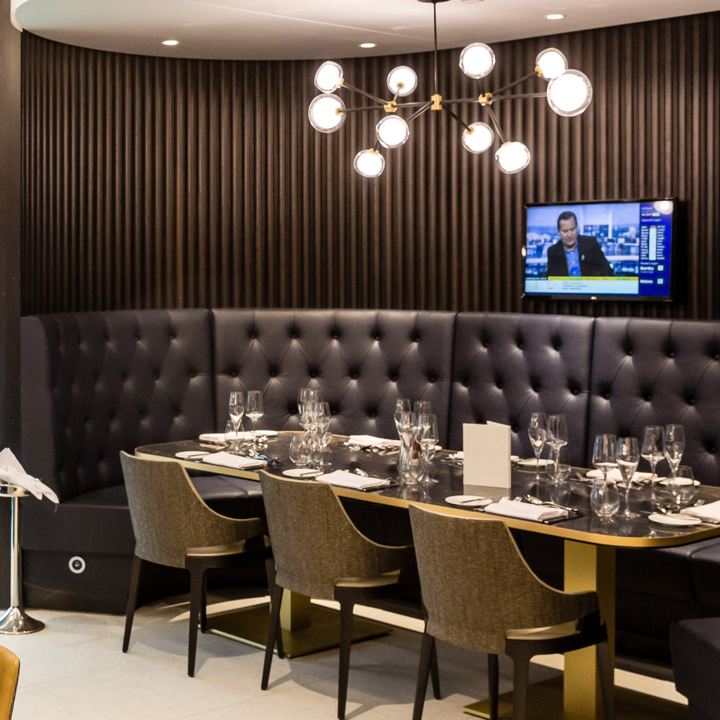 Lit globe chandelier like this Clérachic one, above a long dark dining table with olive chairs, a tufted leather banquette and a wall television.