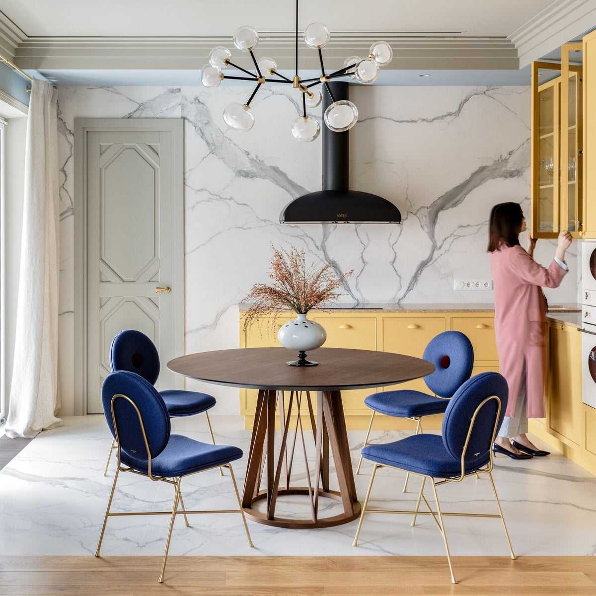 Clérachic chandelier with clear glass globes above a round walnut table with blue chairs, in a marble-walled kitchen with yellow cabinets.