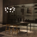 Lit Clérachic chandelier in a dark room, above a black dining table with wooden chairs, in front of a perforated metal screen.