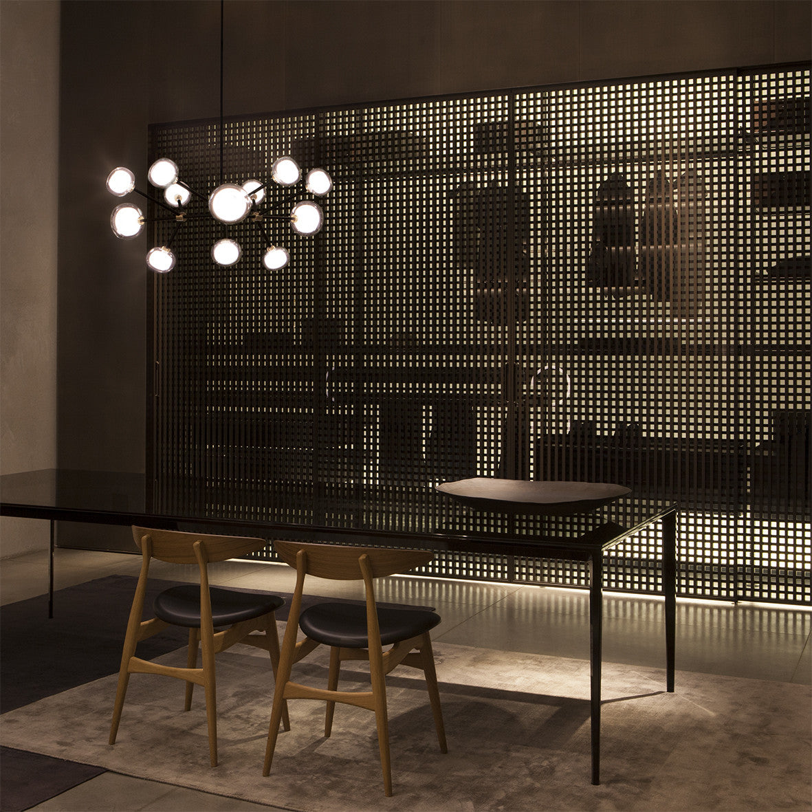 Lit Clérachic chandelier in a dark room, above a black dining table with wooden chairs, in front of a perforated metal screen.