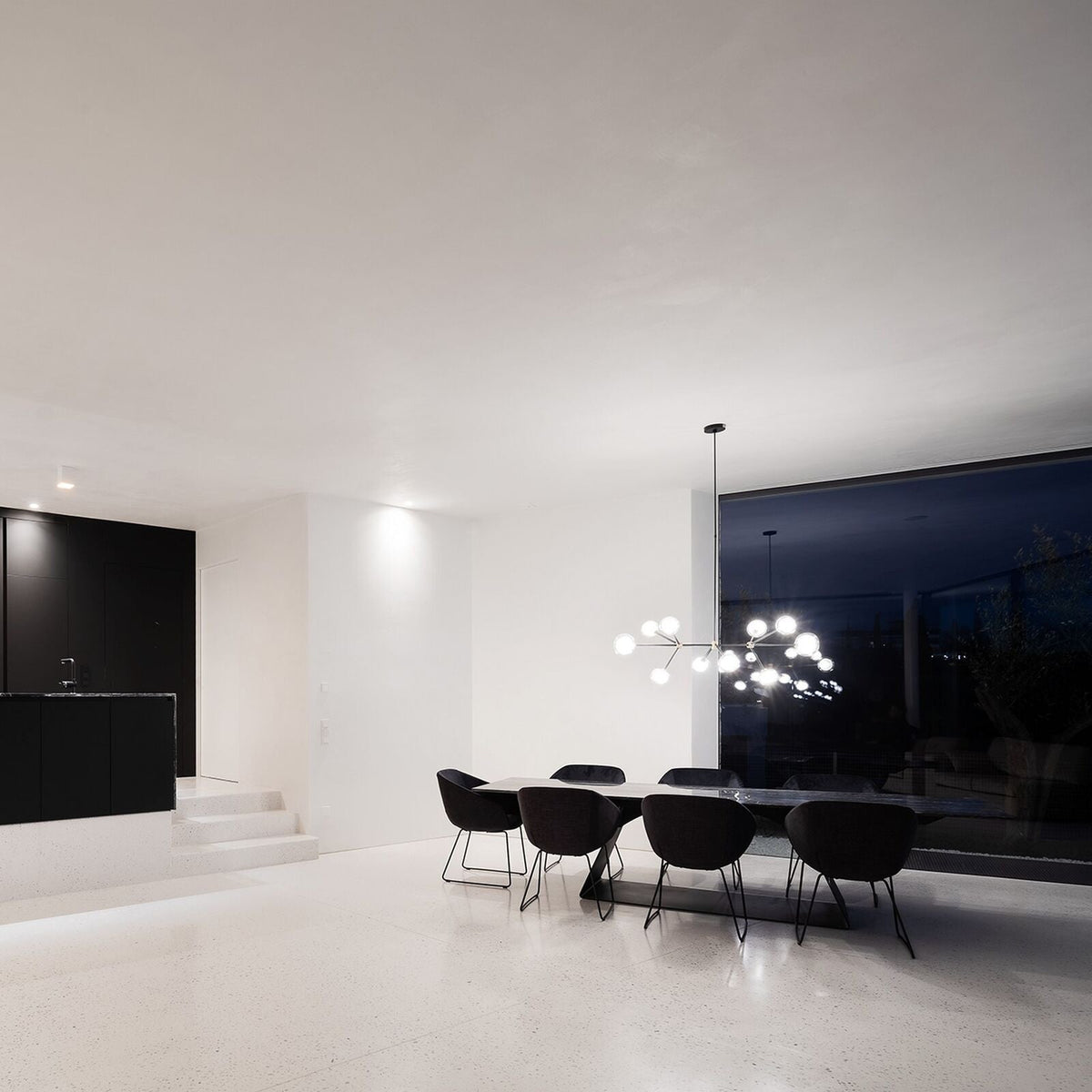 Small Clérachic chandelier lit in the distance above a long dark table with black chairs, in a white room with large dark windows.