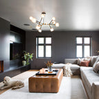 Clérachic chandelier lit with branching arms, in a dark grey living room with a tan leather ottoman, a corner sofa and a small white dog.