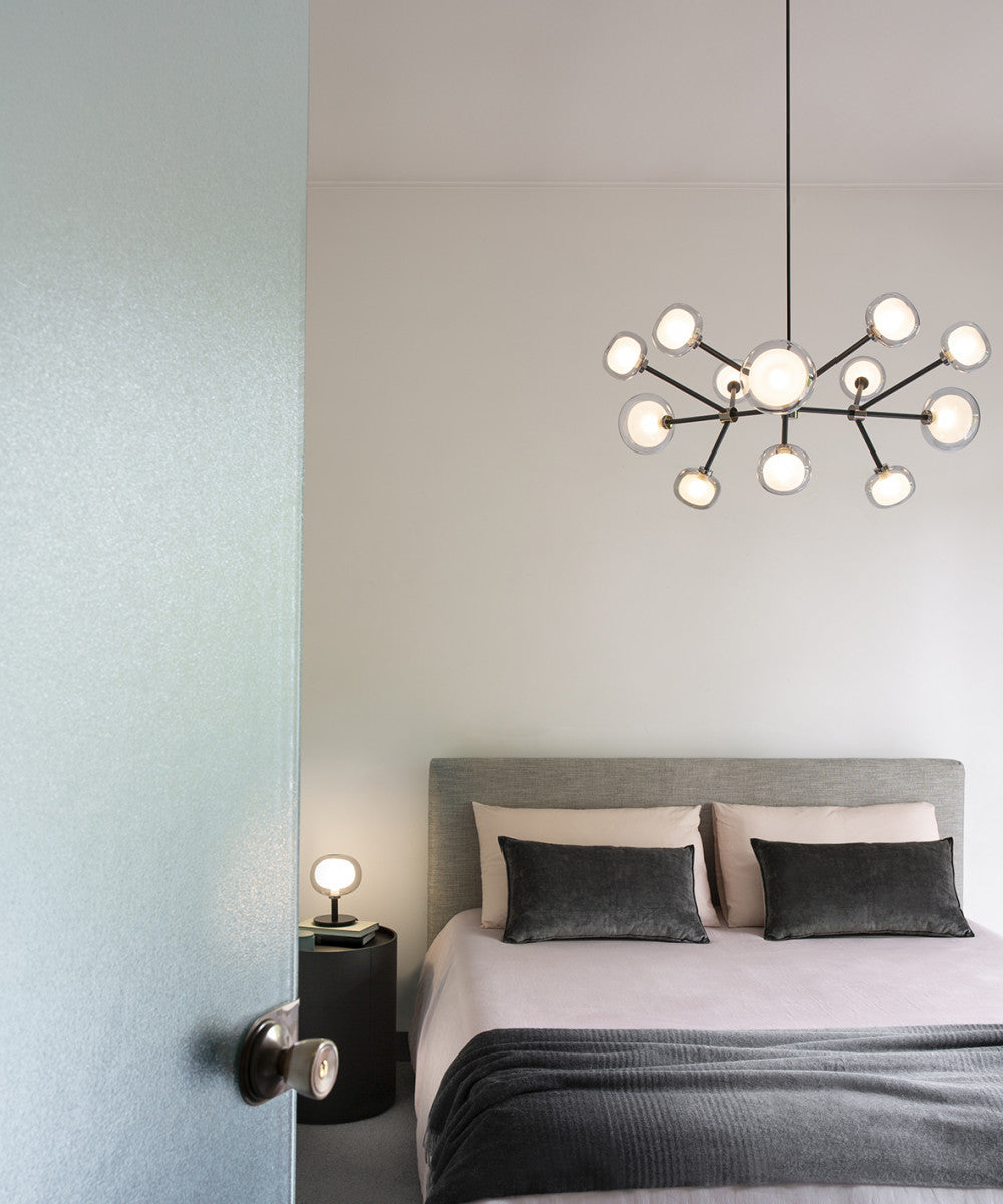 Lit Clérachic chandelier above a bed with grey headboard and dark cushions, seen through a doorway in a pale bedroom.