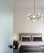 Lit Clérachic chandelier above a bed with grey headboard and dark cushions, seen through a doorway in a pale bedroom.