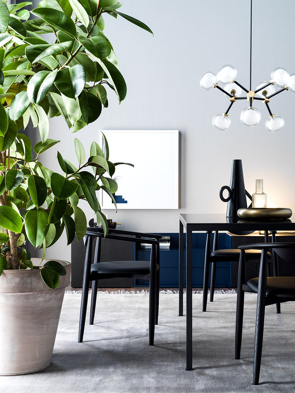 Clérachic chandelier with clear globes beside a large leafy plant, above a black dining table and chairs against a grey wall.