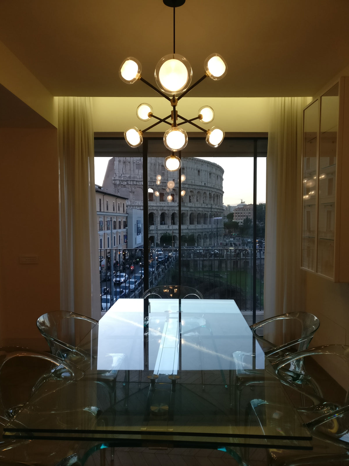 A Clérachic chandelier with a large central globe in a room with a glass dining table, and a view of an ancient amphitheatre through the window.