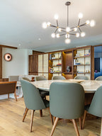 Clérachic chandelier lit above a round marble dining table with grey-green leather chairs, in a room with a wood-framed bookshelf.