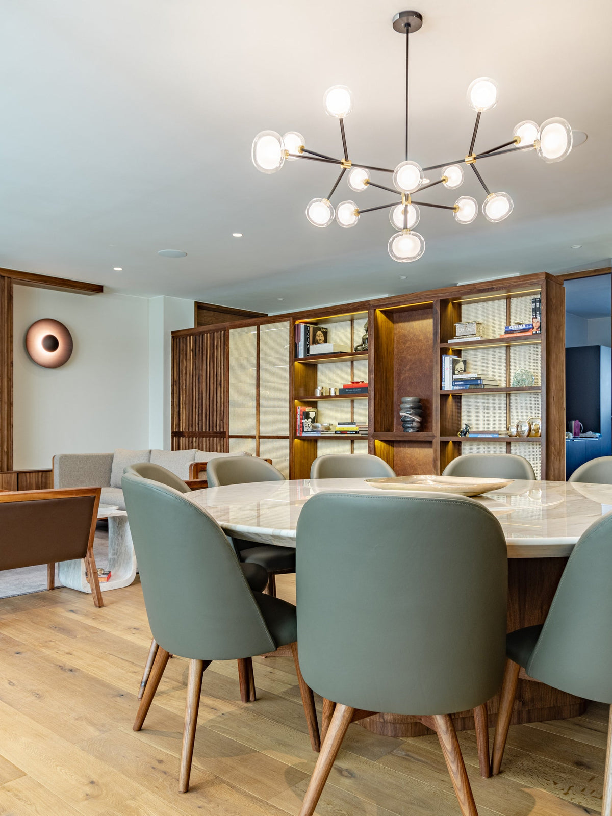 Clérachic chandelier lit above a round marble dining table with grey-green leather chairs, in a room with a wood-framed bookshelf.