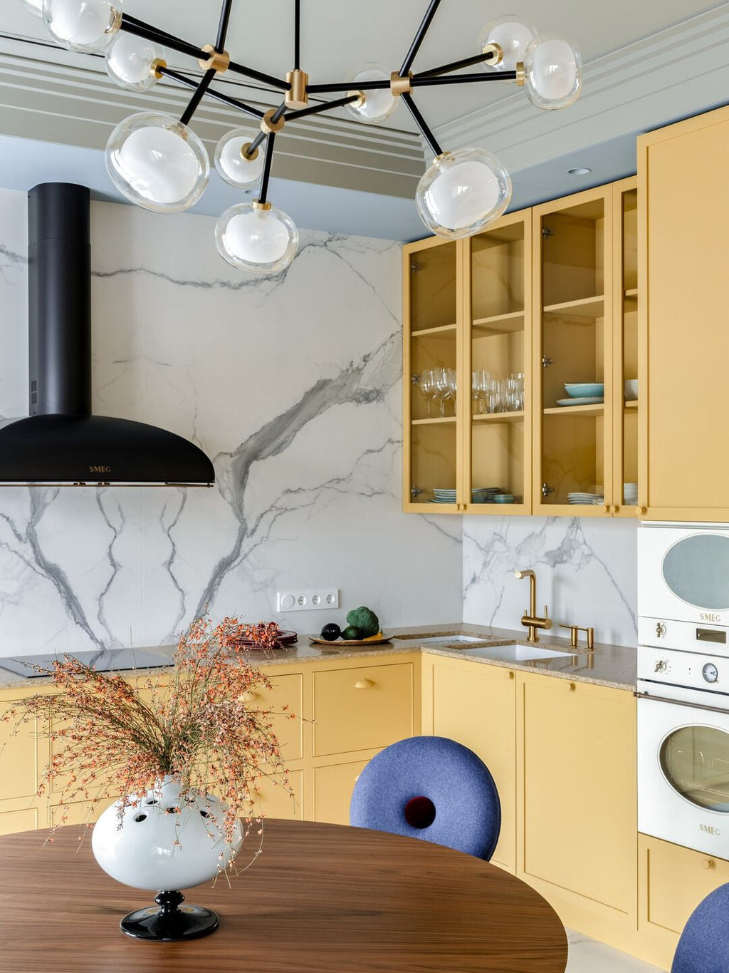 Clérachic chandelier with clear globes above a walnut table and a vase of dried stems, in a marble-backed kitchen with yellow cabinets.