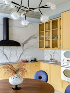 Clérachic chandelier with clear globes above a walnut table and a vase of dried stems, in a marble-backed kitchen with yellow cabinets.