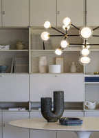 Lit Clérachic chandelier in front of cream shelving with vases and books, above a white oval table with a dark ceramic vase.