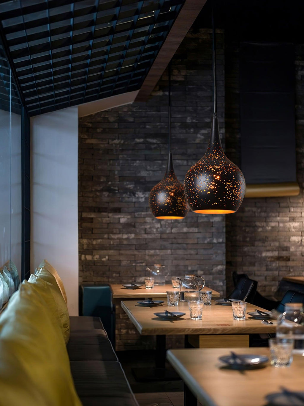 Two small teardrop Clérachic pendants hanging in a restaurant with a dark brick wall, wooden tables and a mustard banquette.