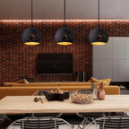 Three round Clérachic pendants in a line above a long pale table, in front of a red brick wall with a television and a mustard sofa.