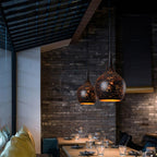 Two teardrop Clérachic pendants lit in a restaurant corner, with dark brick walls, a slatted ceiling and laid tables below.