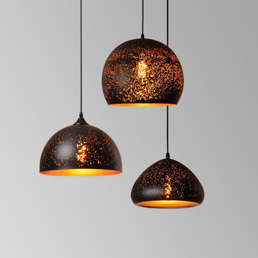 Three Clérachic pendants in dome and round shapes hanging at different heights against a light grey wall, their gold interiors glowing.