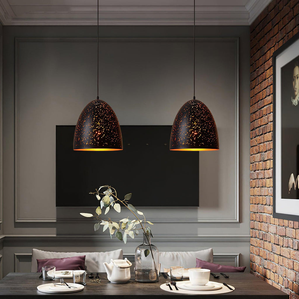 Two Clérachic bell pendants above a dark dining table, with a brick wall, a wall-mounted television and a vase of leaves behind.