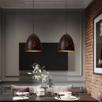 Two Clérachic bell pendants above a dark dining table, with a brick wall, a wall-mounted television and a vase of leaves behind.