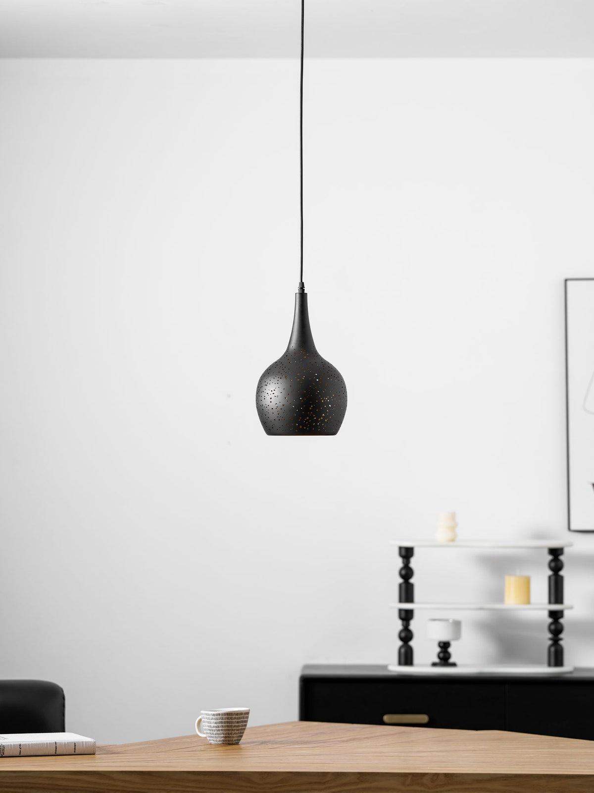 Small teardrop Clérachic pendant on a long black cord, above a wooden table with a cup, in front of a white wall and black sideboard.