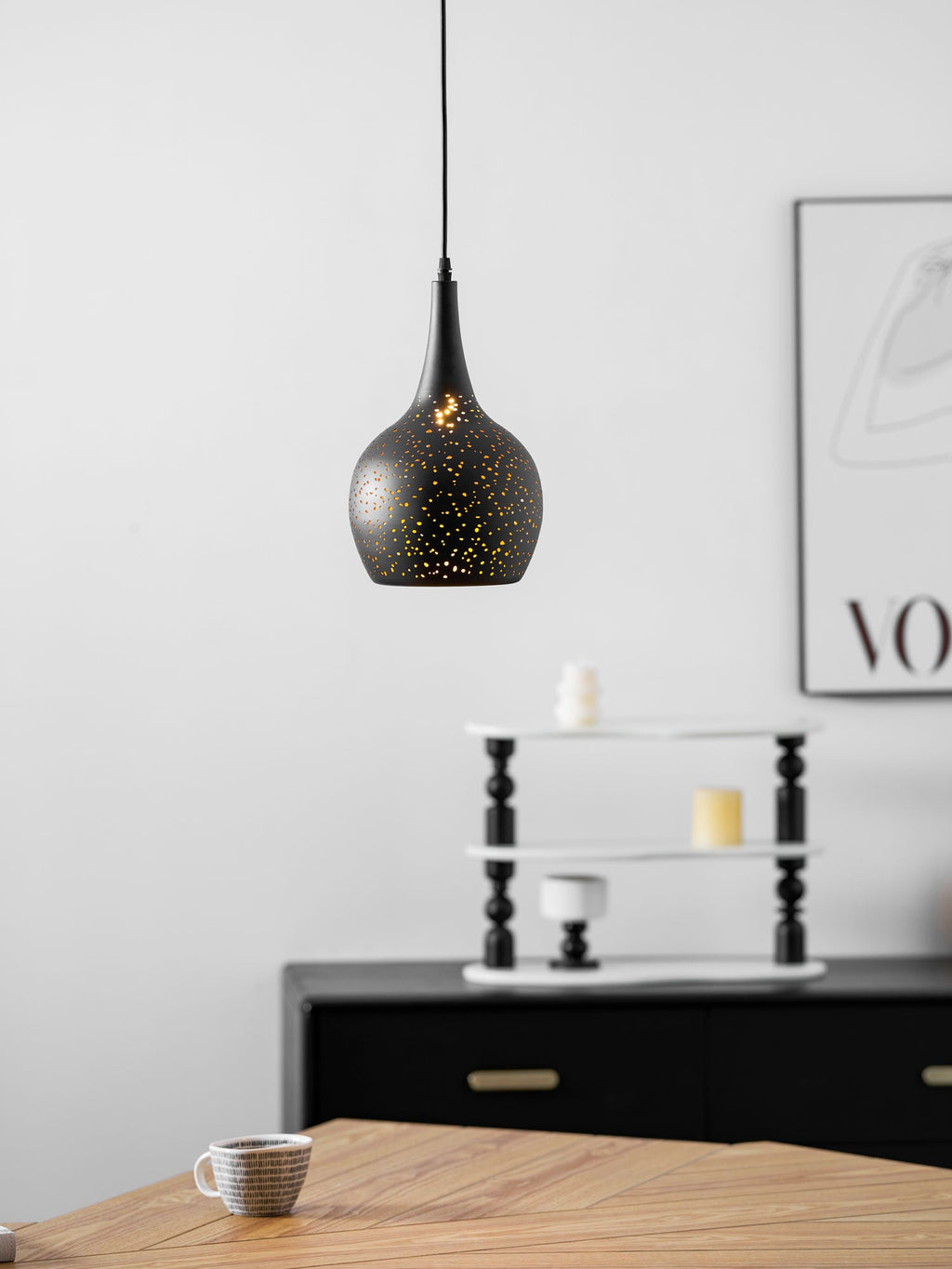 Closer view of the black teardrop Clérachic pendant, with a framed print and a stacked shelf behind and a cup on the table below.
