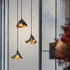 Three Clérachic leaf-shaped pendants hang at staggered heights against a pale window with misty hills and red blossoms.