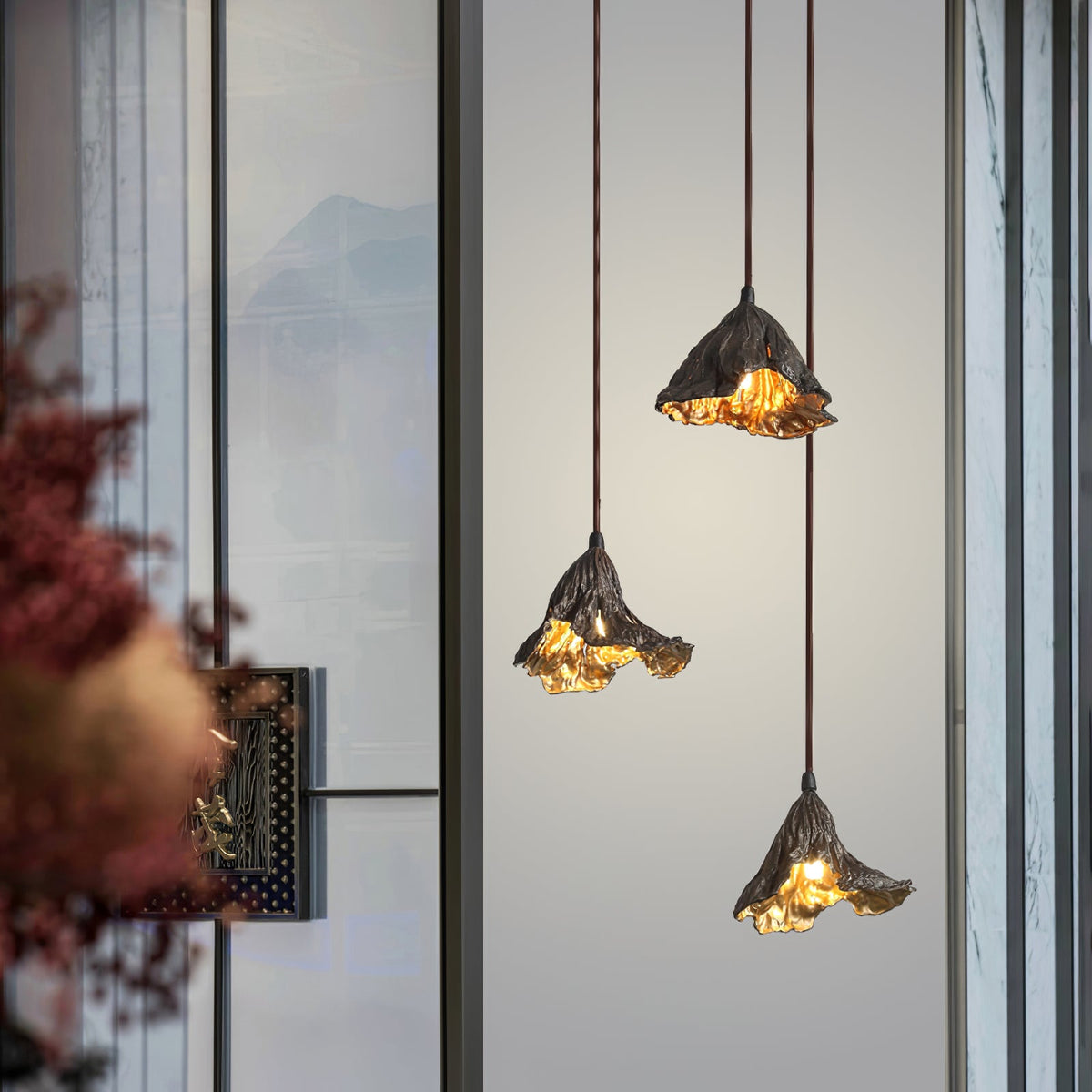 This Clérachic pendant trio in black and gold hangs in front of a glazed window, with red autumn foliage at the left edge.