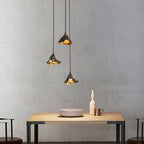 Three Clérachic pendants of black metal with gold inside hang over a pale wood table, against a rough grey plaster wall.