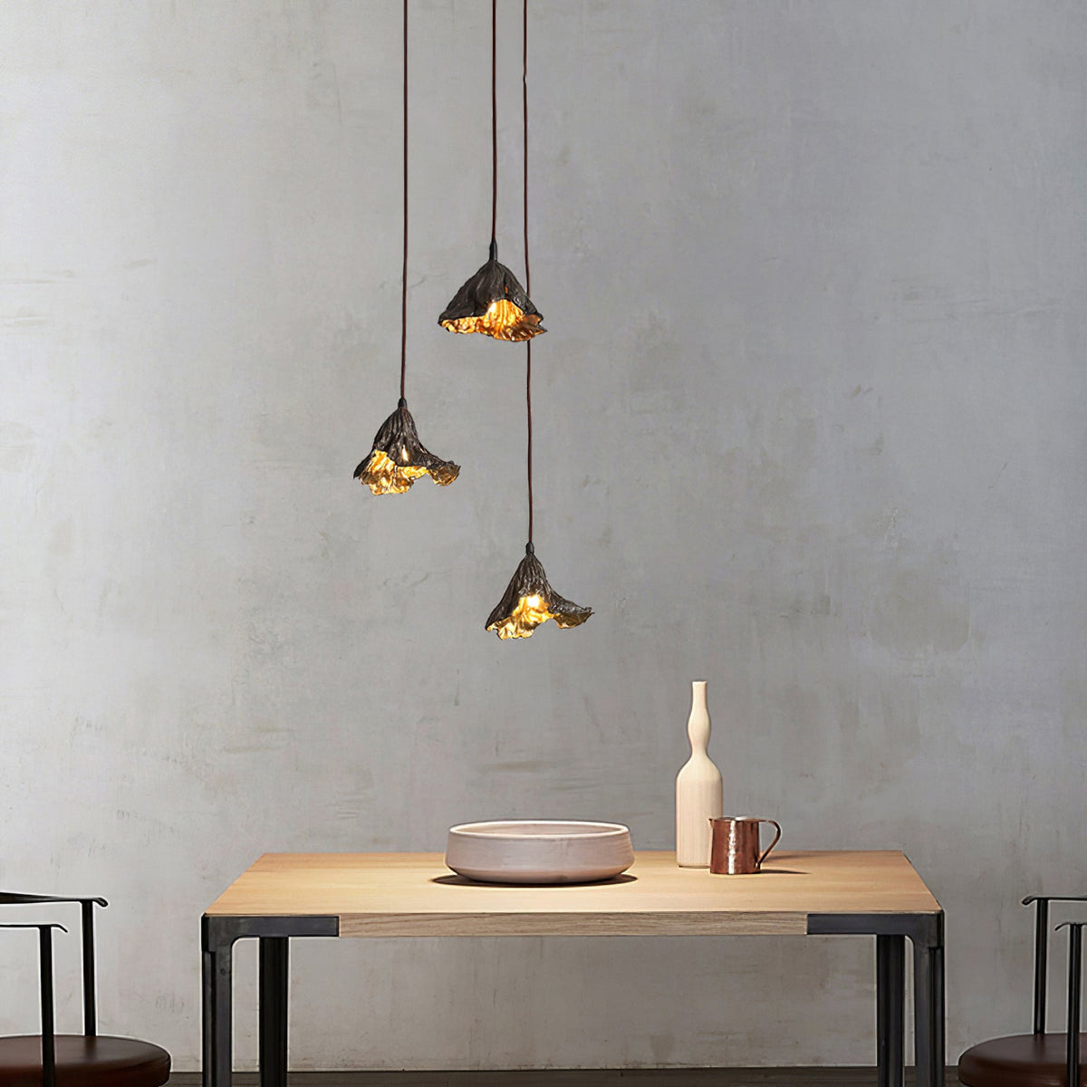 Three Clérachic pendants of black metal with gold inside hang over a pale wood table, against a rough grey plaster wall.
