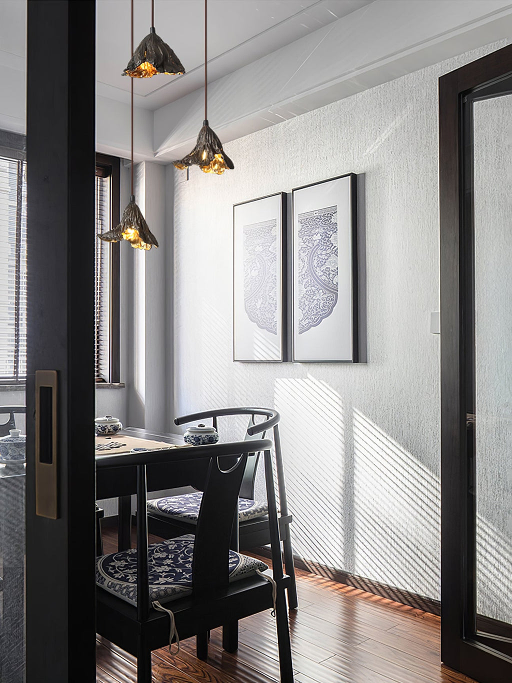 Two Clérachic pendants hang in a dining nook with dark chairs, two framed prints and sunlight across a textured wall.