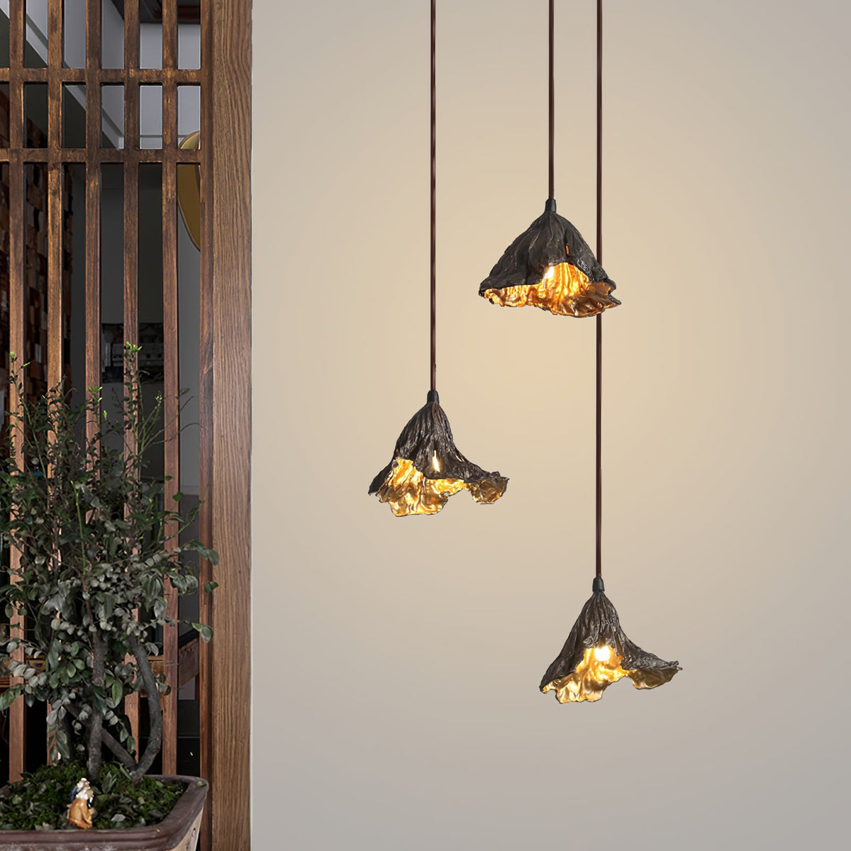 Three Clérachic pendants hang in front of a wood-slatted partition and a potted plant, lit warm against a beige wall.