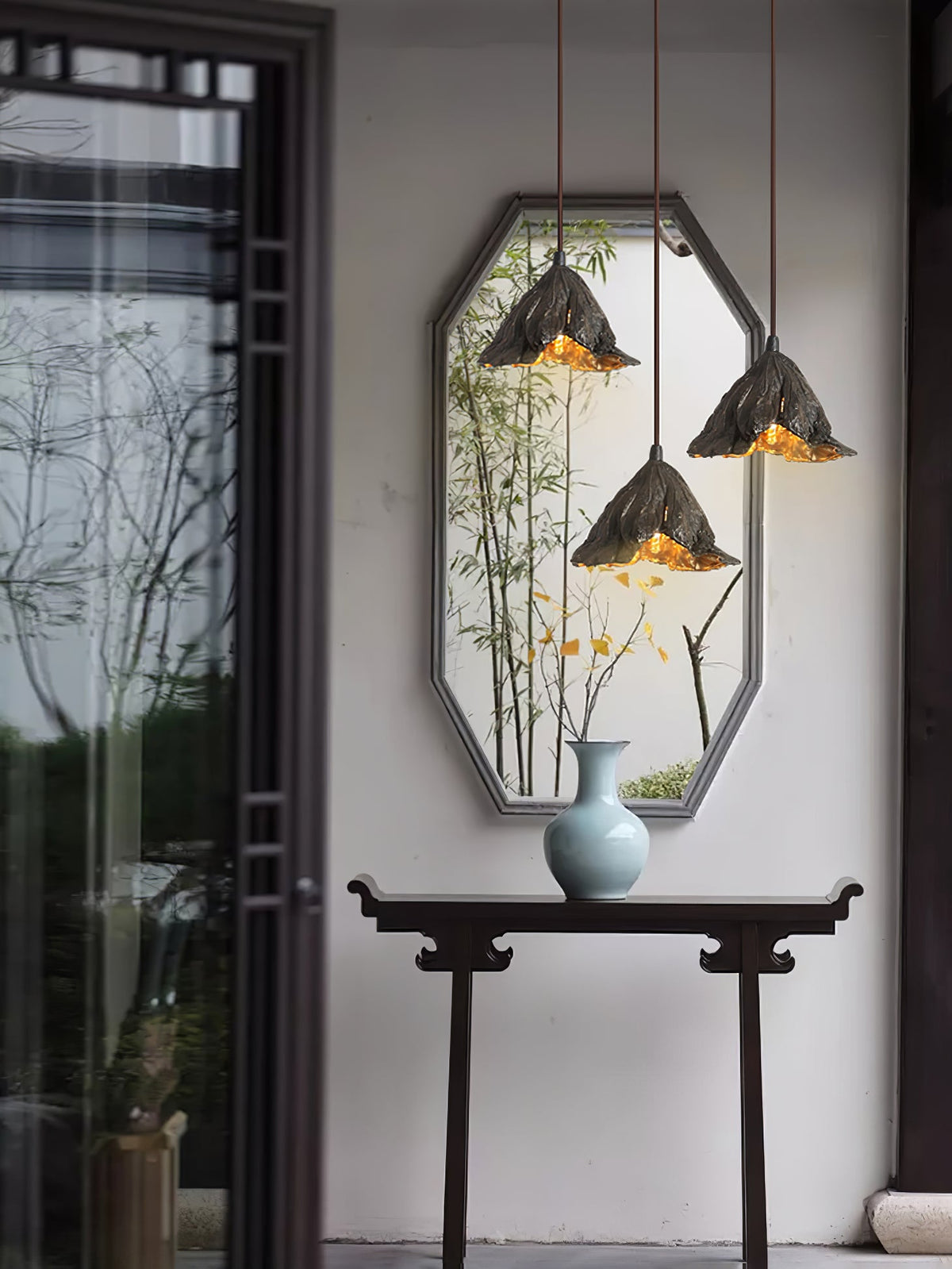 Three Clérachic pendants hang in front of an octagonal mirror with bamboo, above a dark console table and a pale blue vase.