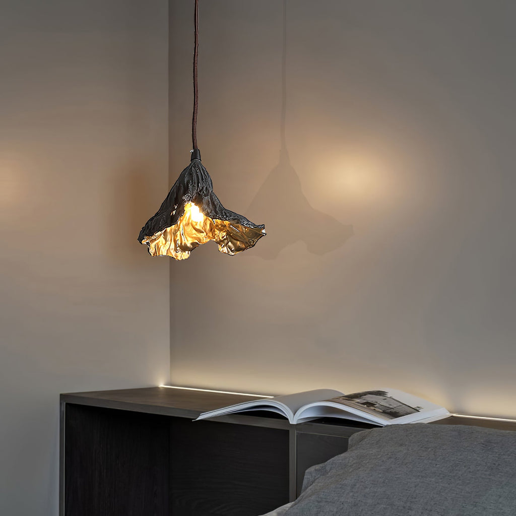 A Clérachic pendant lit warm in a bedroom corner, above a dark shelf with an open magazine and a grey bed.