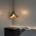 A Clérachic pendant lit warm in a bedroom corner, above a dark shelf with an open magazine and a grey bed.