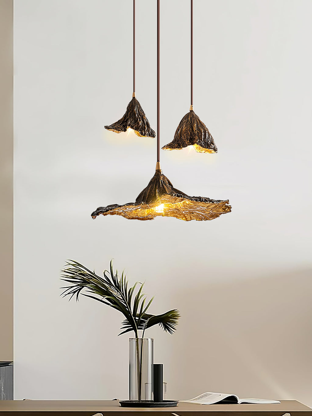 Three Clérachic pendants hang in a pale room, the lowest with a wide flat shade, above a vase of palm leaves on a table.