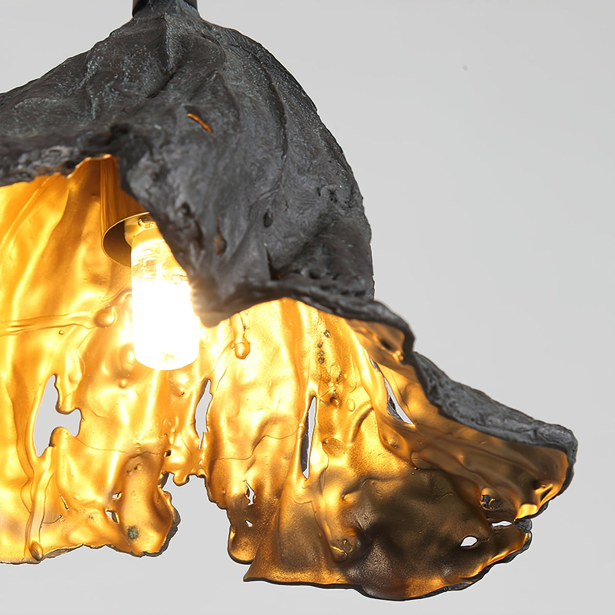 Close view of the bulb inside this Clérachic pendant, lit within a gold interior with cut-out openings and a rough black shell.