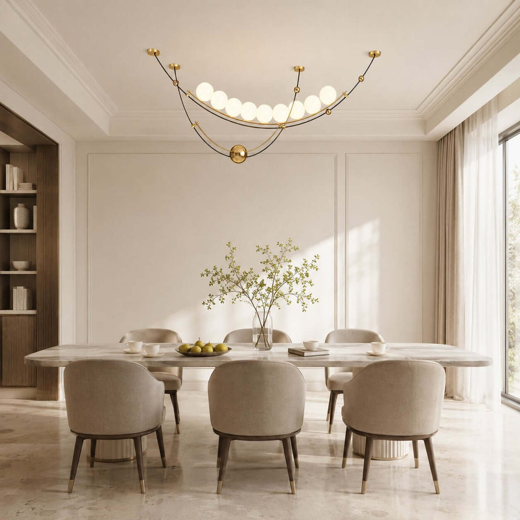 A Clérachic pendant with a row of glowing globes above a long marble dining table with six upholstered chairs in a pale room.