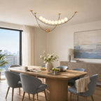Lit Clérachic pendant with curved black and gold arcs over an oak dining table, with blue-grey chairs and a window onto the city.