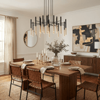 A Clérachic tubular chandelier hangs over a candle-lit dining table, with woven chairs and a bold black and gold painting behind.