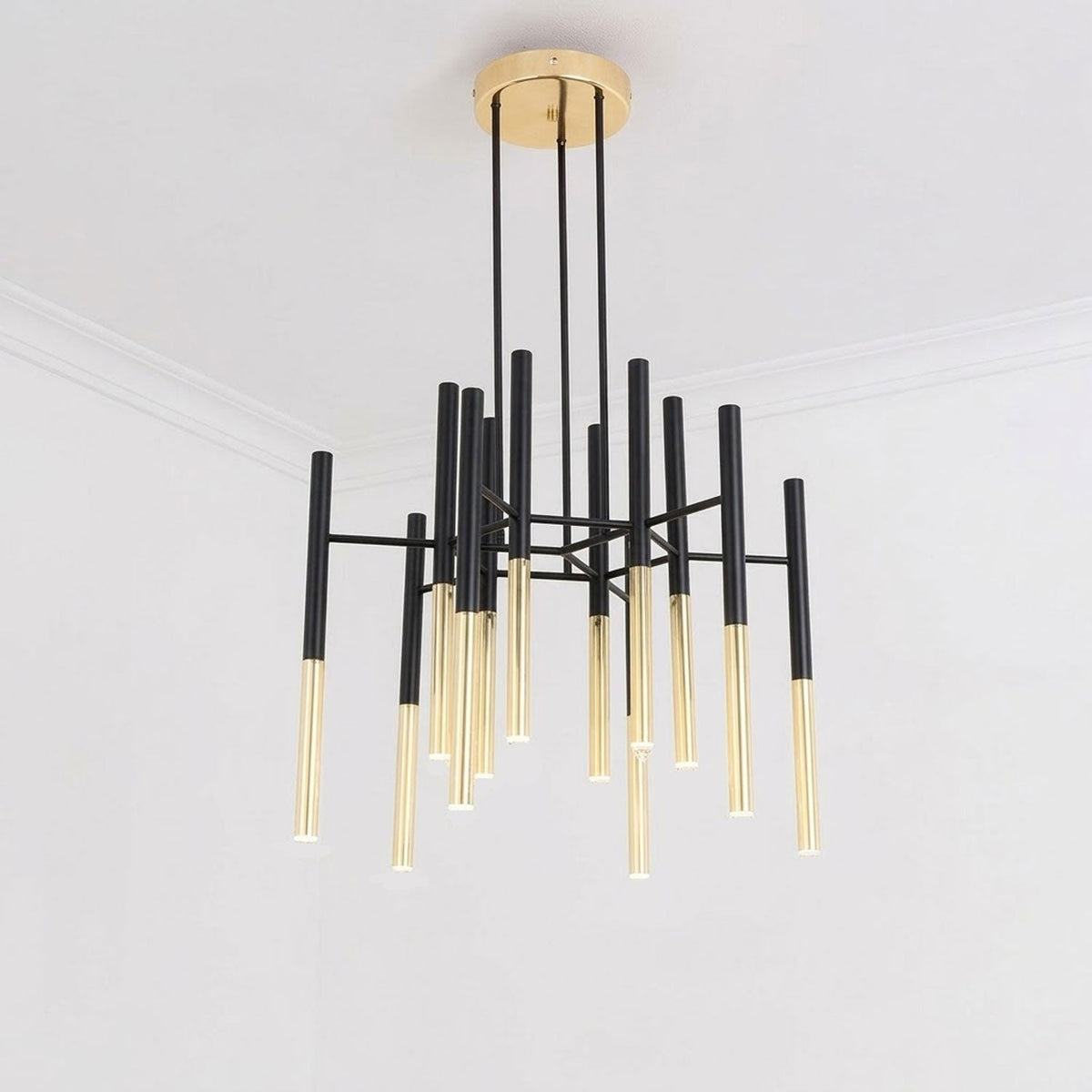 A Clérachic chandelier with black arms and gold-finish tubes hangs from the ceiling of a plain white room, seen from below.